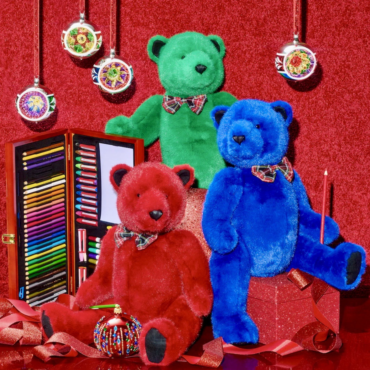 Three plush teddy bears in green, red, and blue colors posed with holiday ornaments and colored pencils