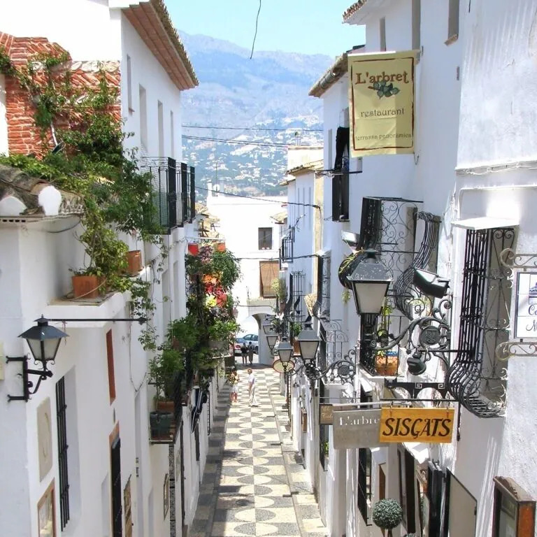 Narrow cobblestone street between whitewashed buildings in Altea, Spain, with mountains beyond