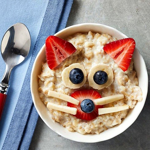 Oatmeal bowl decorated with fruit arranged as a cat face with strawberry ears, banana whiskers, and blueberry eyes