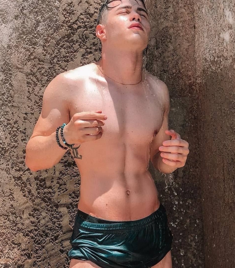 Young muscular man in dark swim briefs showering outdoors against a stone wall