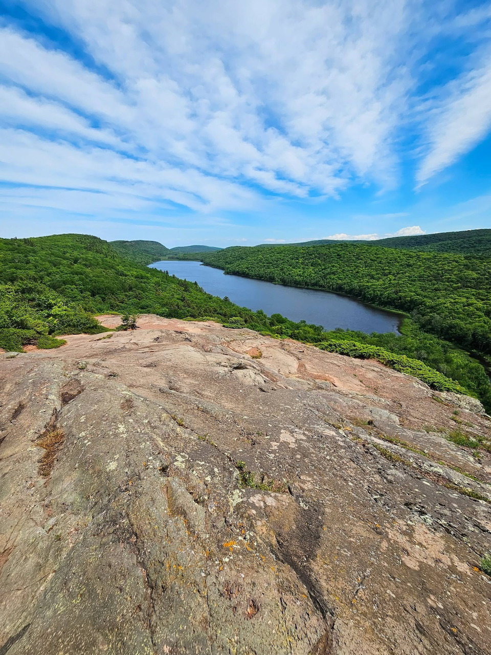 Rocky cliff overlooking forested lake and mountains under blue sky with clouds