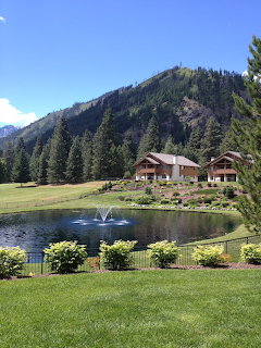 Wooden lodge buildings near a pond with fountain surrounded by green lawns and forested mountains
