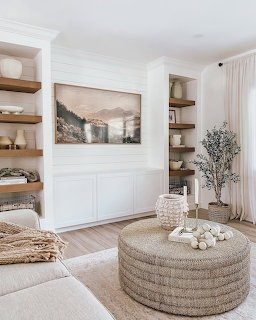 Neutral-toned living room with white built-ins, wooden shelves, woven ottoman, and framed landscape art