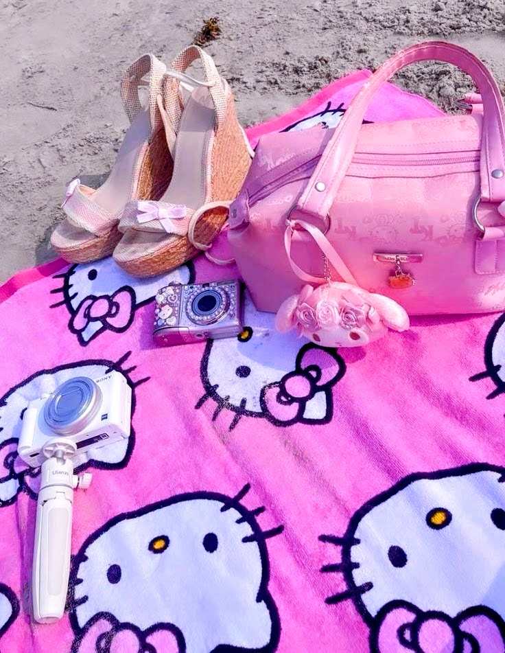 Pink Hello Kitty beach towel on sand with pink bag, wedge shoes, camera, and accessories