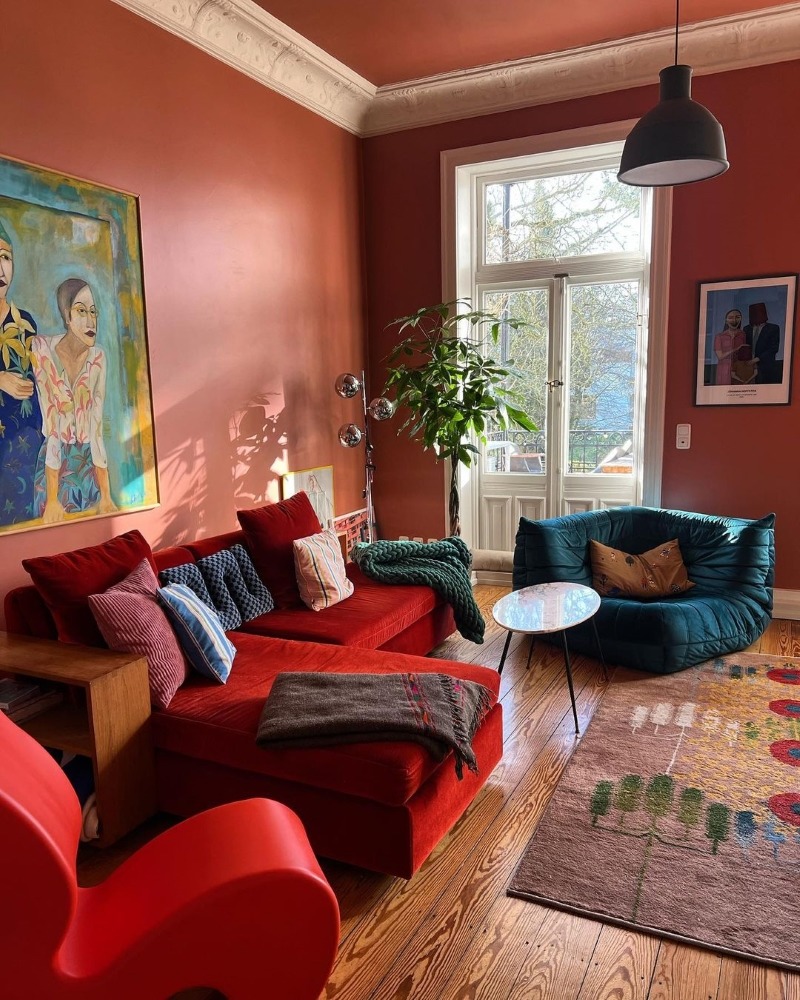 Cozy living room with red walls, colorful sofas, artwork, window, and wooden floor