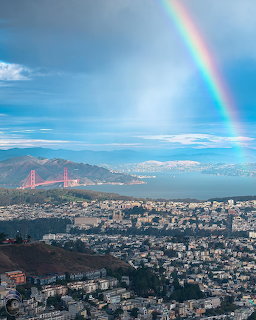 Rainbow arcs over a city with red bridge, buildings, and mountains under blue sky