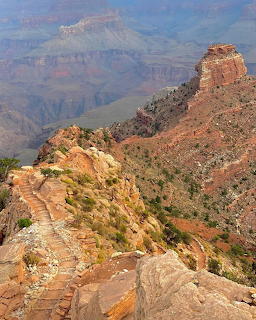Rocky trail along a steep canyon rim with red cliffs and a hazy gorge beyond
