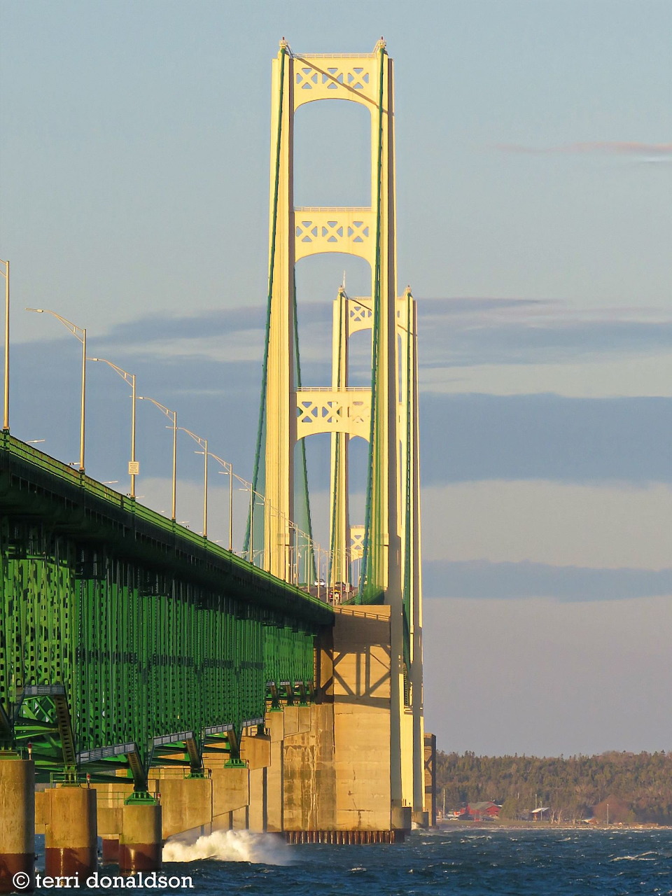 Sunlit Mackinac Bridge towers above blue water with green steel truss and soft clouds