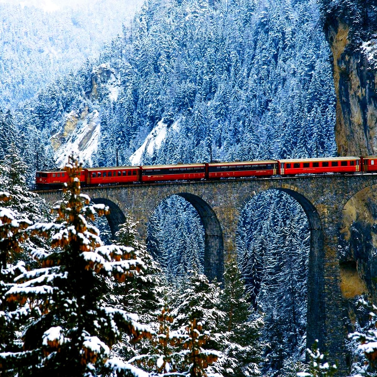 Red train crossing stone bridge through snowy mountain forest with white-frosted trees