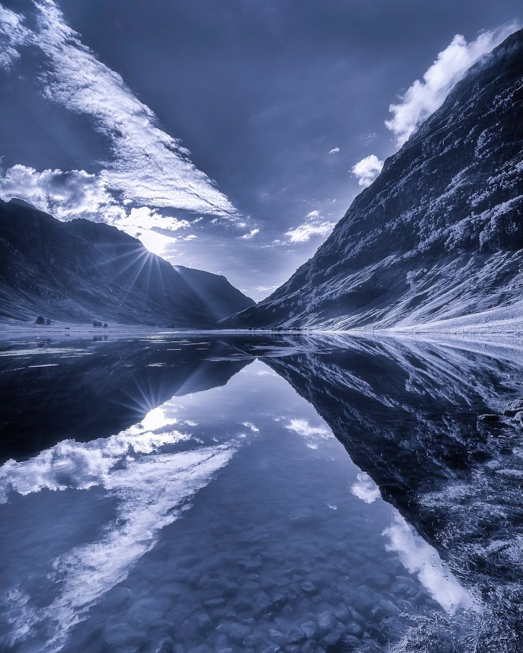 Steep mountains and clouds reflected in a calm alpine lake in blue monochrome tones