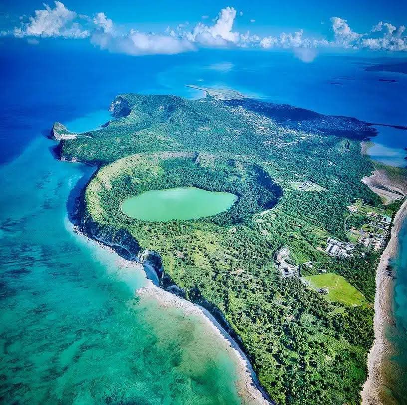 Aerial view of a green island with a circular crater lake and turquoise ocean