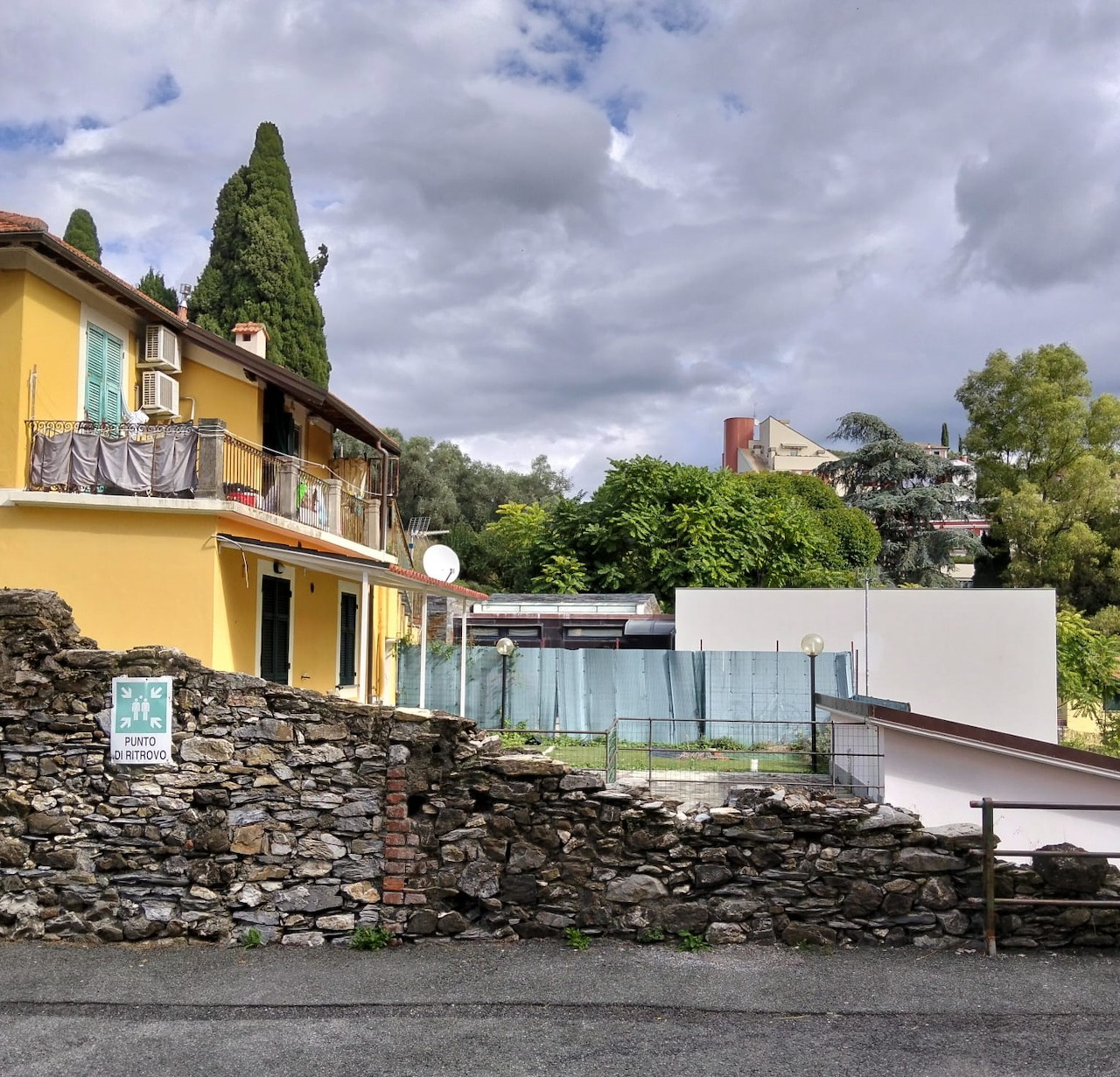 Stone wall ruins beside yellow building with cypress trees and cloudy sky in Italian village