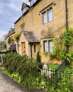 Stone English cottage with ivy, garden plants, and a black iron fence beside a narrow lane