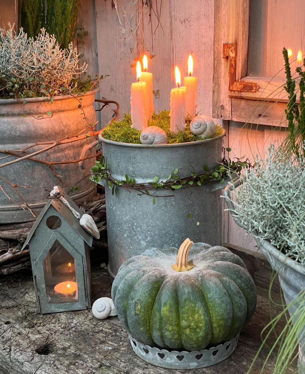 Lit pillar candles in a zinc bucket beside a green pumpkin, small lantern, shells and moss in an autumn outdoor setting.