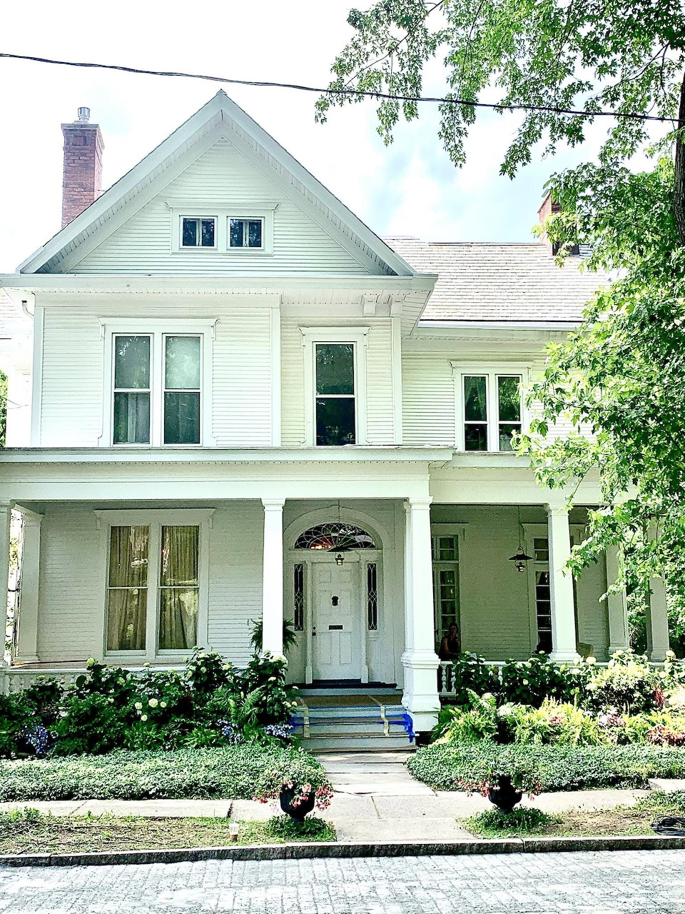 Two-story white Victorian house with front porch, arched doorway, and green trees and garden