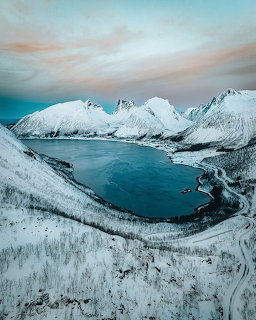 Aerial photo of a snow-covered fjord with jagged mountains and a dark blue bay under a colorful winter sky