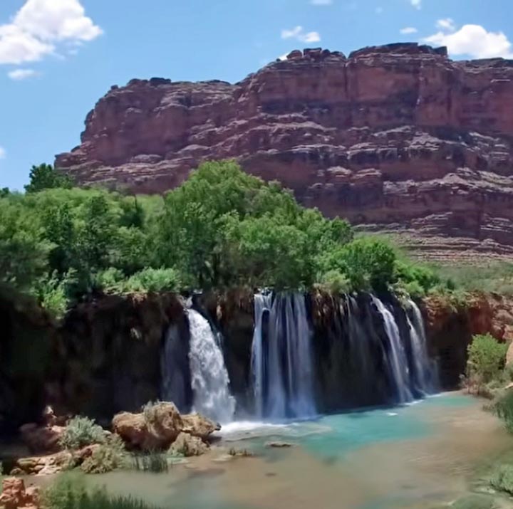 Waterfall flowing into turquoise pool with green trees and red rock cliffs in background