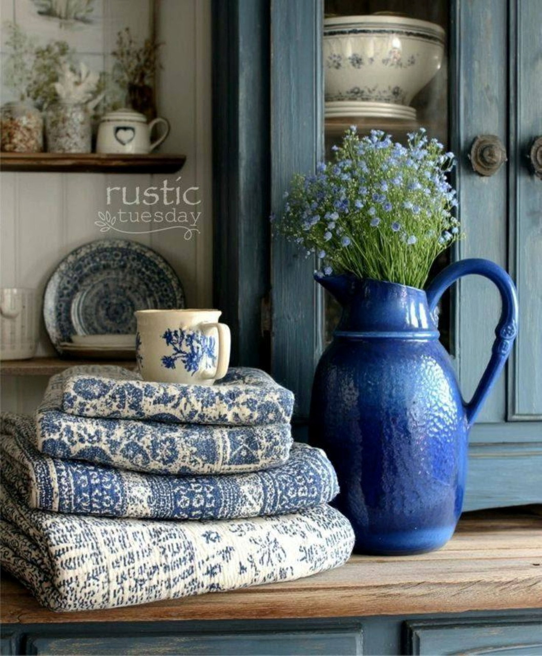Blue rustic home decor with a ceramic pitcher of flowers, folded linens, and vintage dishes on a shelf