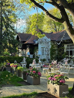 Garden patio with white chairs, flower planters, and statues in front of a cottage-style building under green trees