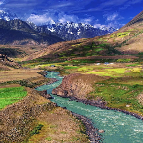 Aerial view of turquoise river flowing through mountain valley with green fields and snow-capped peaks