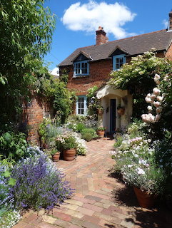 Red brick English cottage with flowering garden, climbing roses, and brick pathway on a sunny day
