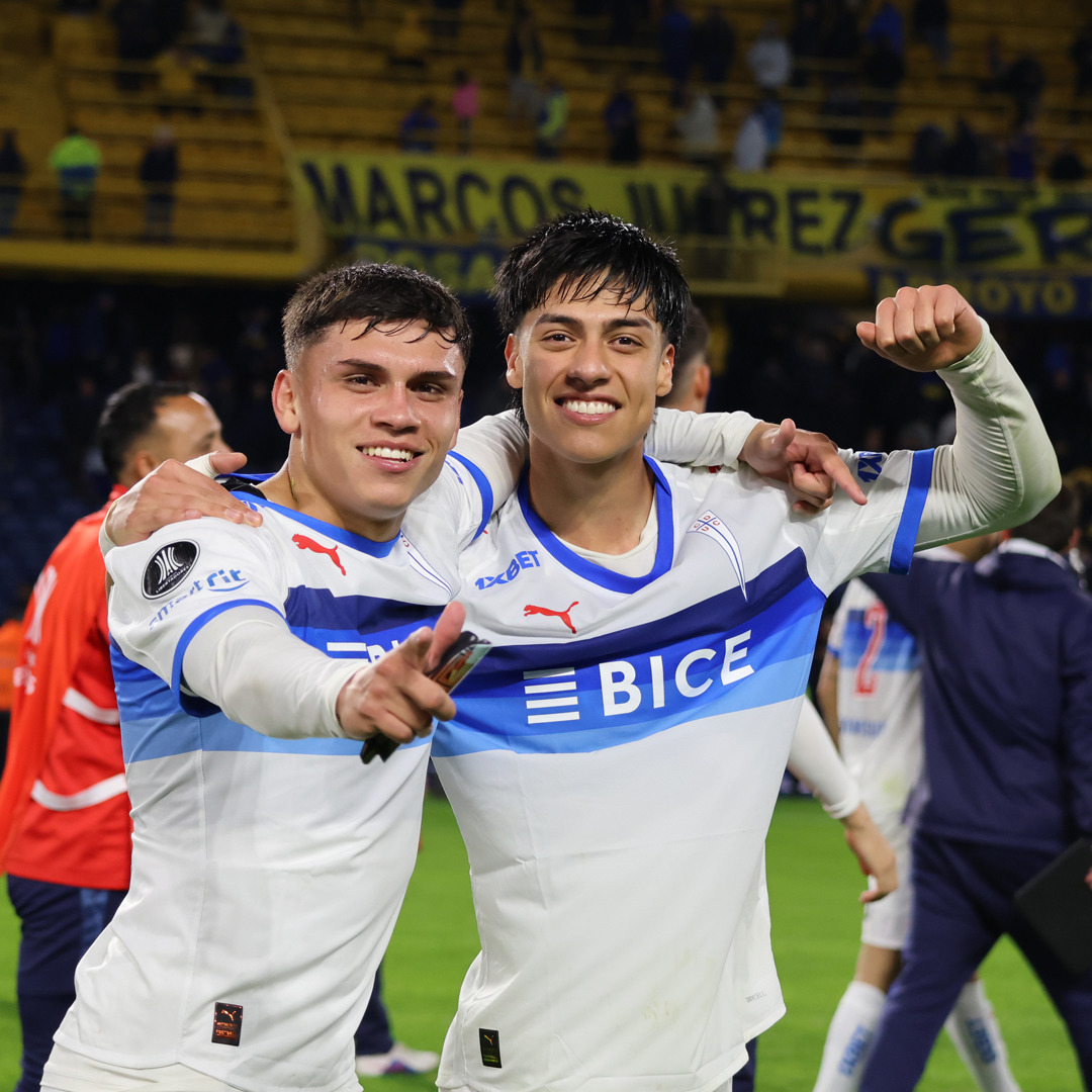 Two smiling soccer players in white jerseys embracing and celebrating on a stadium field