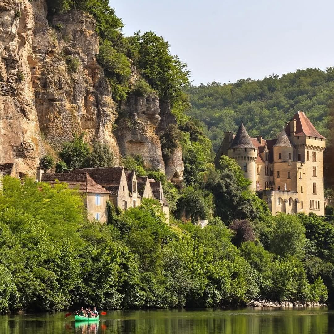 Stone castle and village houses below a rocky cliff beside a calm river and dense trees