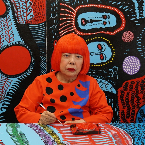 Artist with red bob wig wearing orange polka dot top painting at desk against colorful patterned backdrop