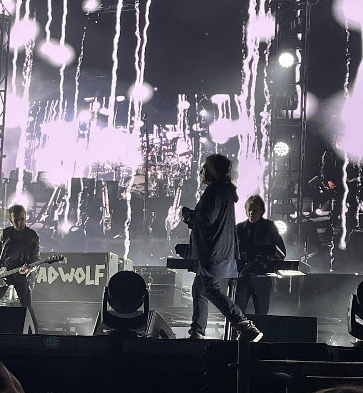 Band members of The Cure performing on stage with white splatter visuals on a large screen backdrop
