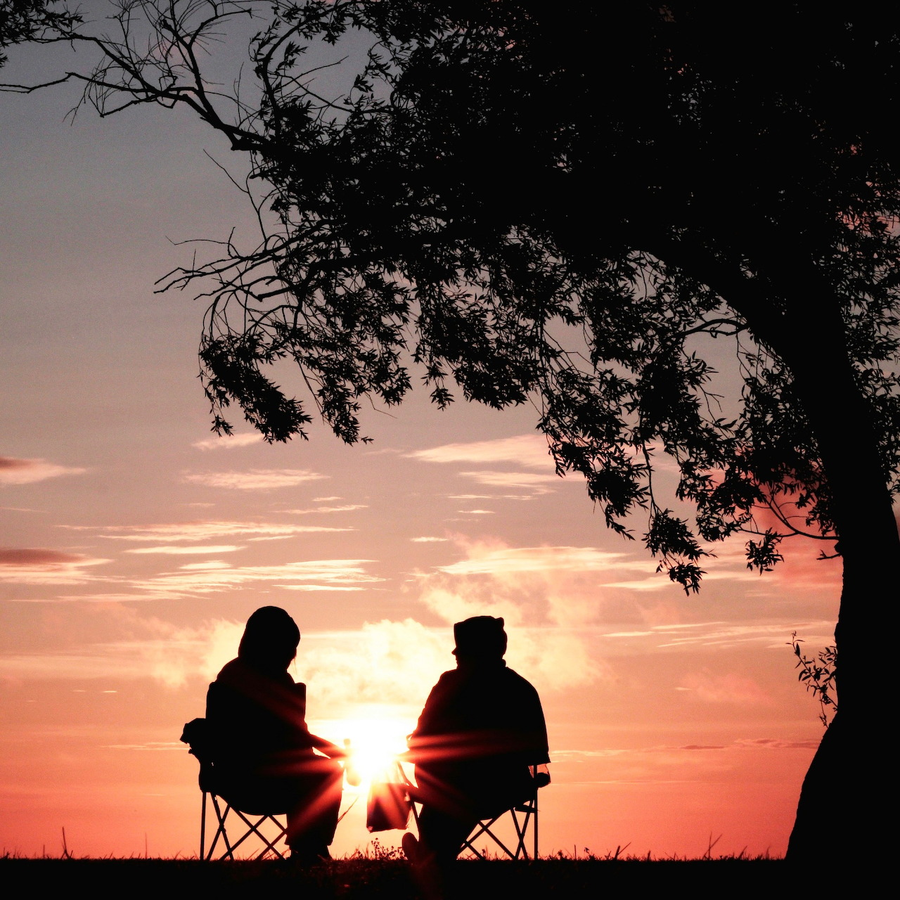 Silhouette of two people sitting in chairs under a tree watching a colorful sunset