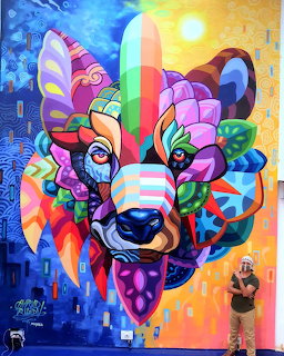 Colorful street art mural of a stylized bear face with geometric patterns and a person standing next to it