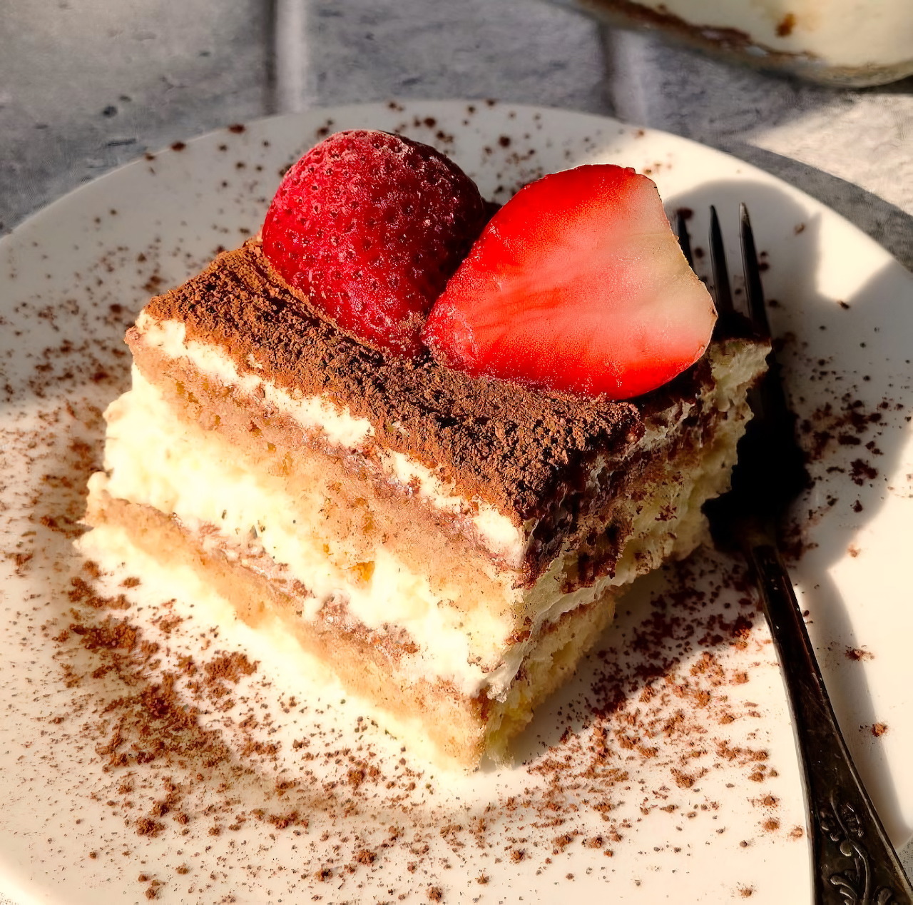 Slice of tiramisu topped with a strawberry on a plate with cocoa powder and a fork