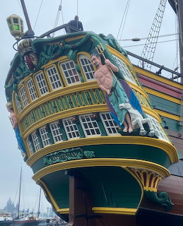 Ornate stern of a historic tall ship with carved figures and green and gold paintwork in a harbor