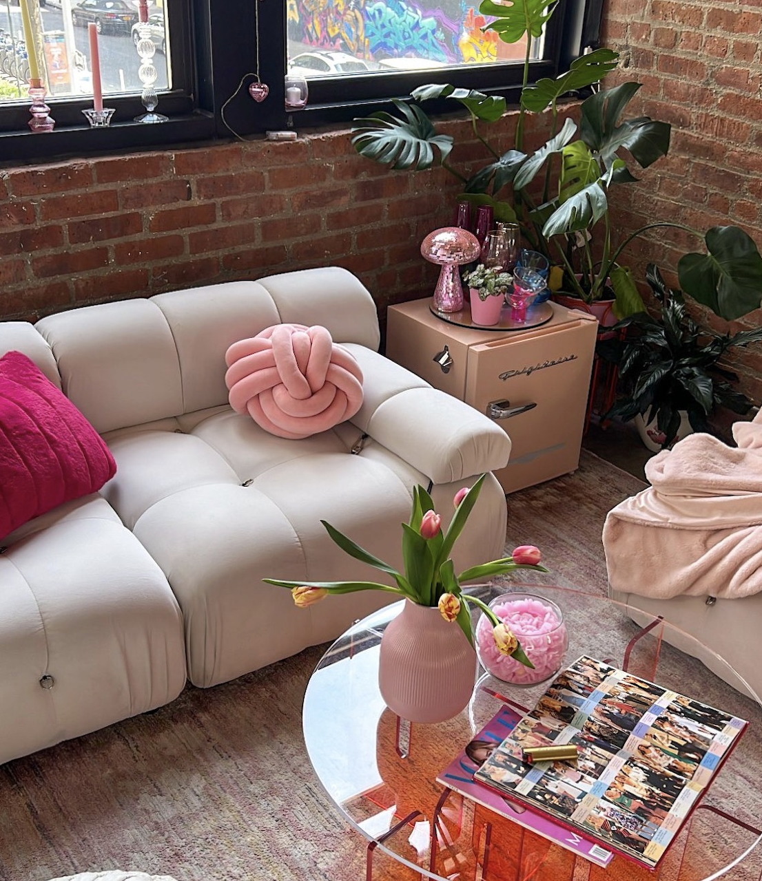 Cozy living room with cream sofa, pink knot pillow, tulips, houseplants, and exposed brick wall