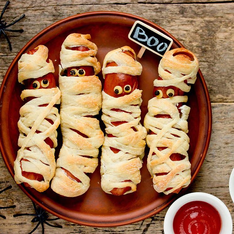 Four mummy-style hot dogs on a plate with ketchup on a rustic wooden table