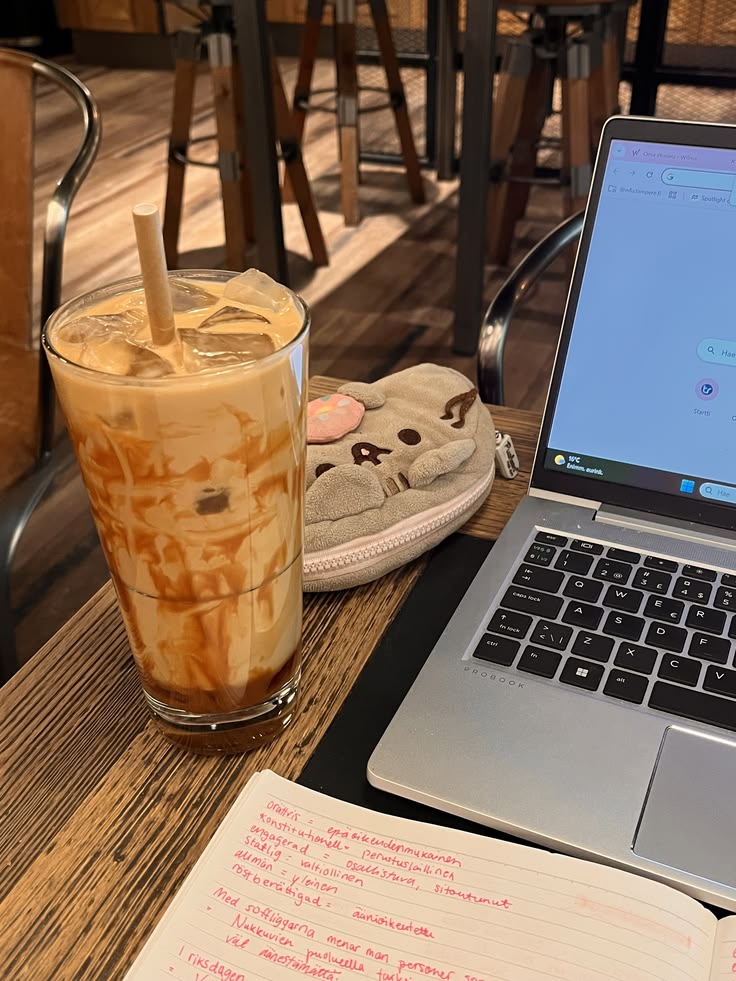 Laptop, iced coffee drink, and handwritten notes on a wooden café table with chairs in background