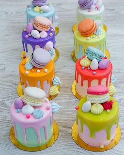 Eight colorful mini drip cakes topped with macarons and meringues arranged on a gray wooden table