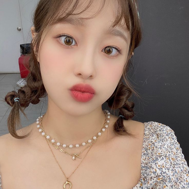 Close-up selfie of a young woman with braided hair, red lipstick, and pearl necklaces indoors