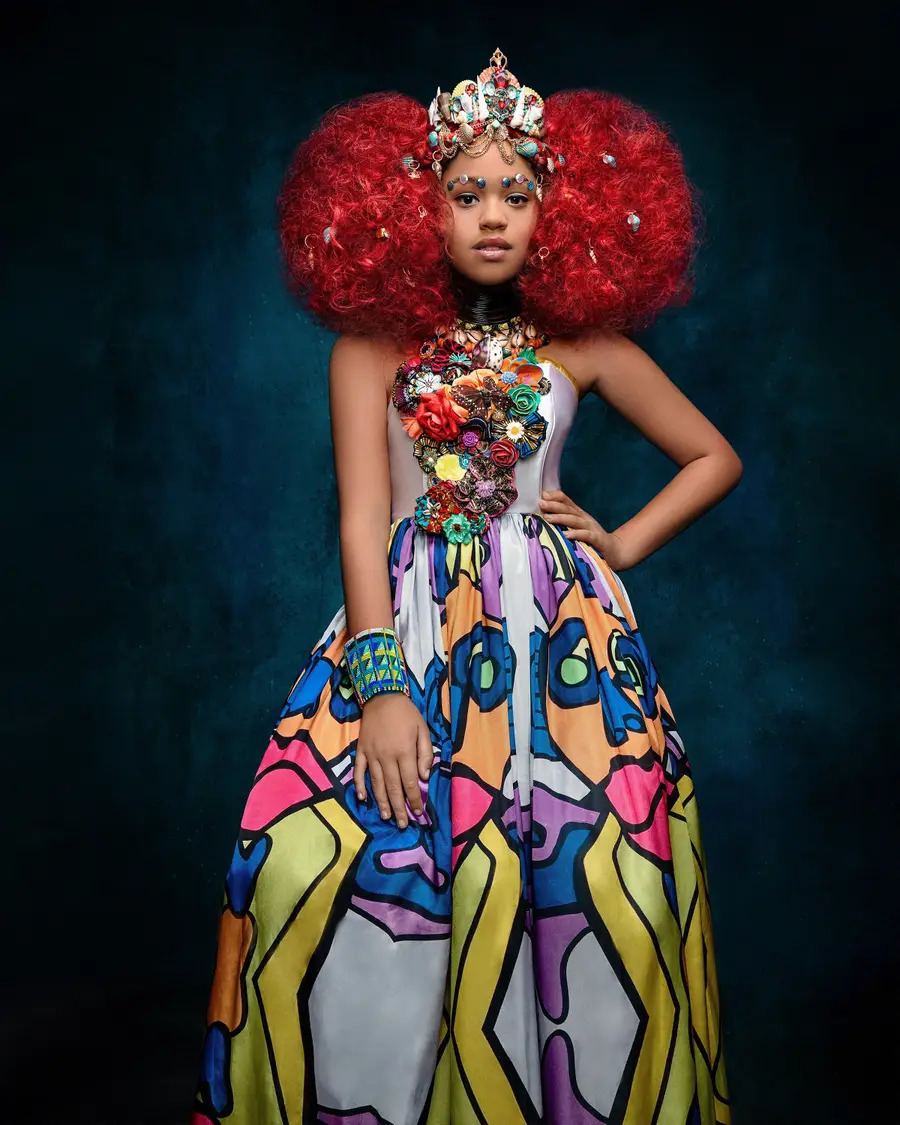 Model in colorful patterned gown with red voluminous hair and ornate crown on dark background