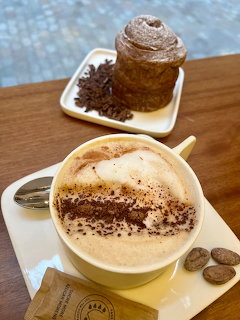 Foamy cappuccino on a wooden table with coffee beans and a pastry behind it