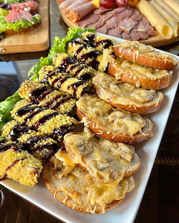 White platter with cheesy open-faced toasts and dark drizzle, charcuterie board with meats and cheese in background