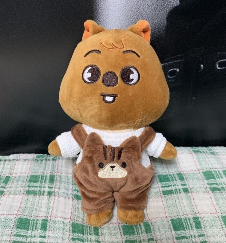 Brown quokka plush toy in white shirt and overalls on green checkered cloth