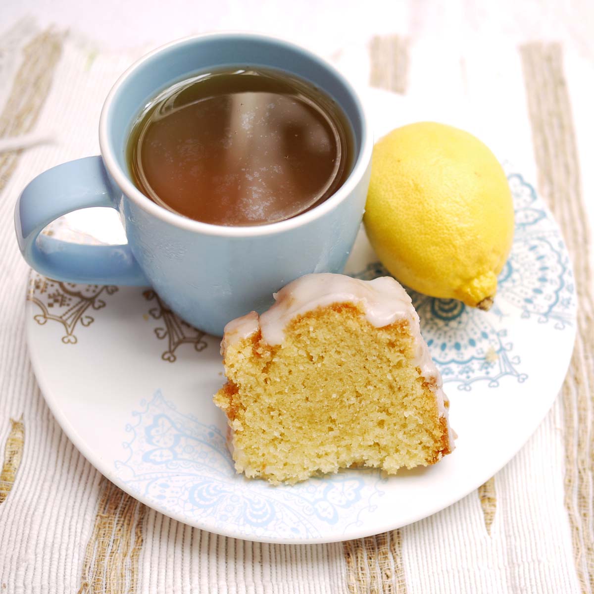 Slice of lemon cake with icing, blue tea cup, and fresh lemon on white plate