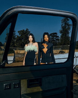 Two women in dark fashion outfits posing outdoors at dusk, viewed through a car window frame