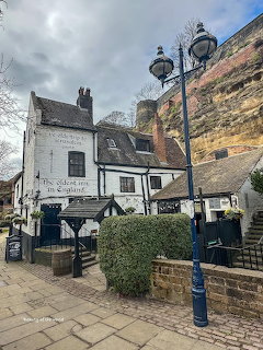 White historic pub building with dark roof nestled against tall sandstone cliff under cloudy sky