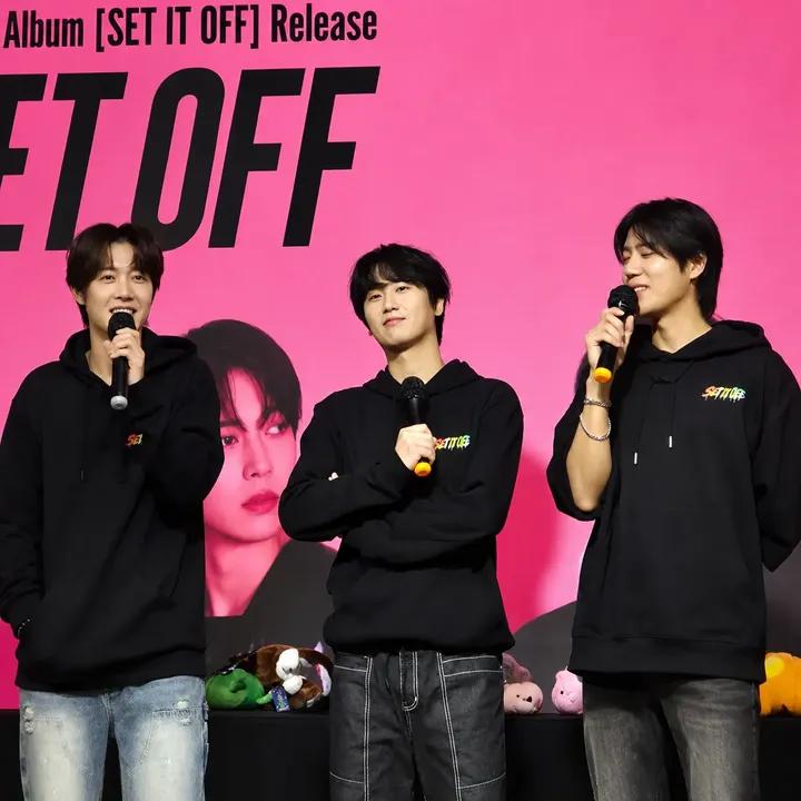 Three young men with microphones on stage before a bright pink promotional backdrop