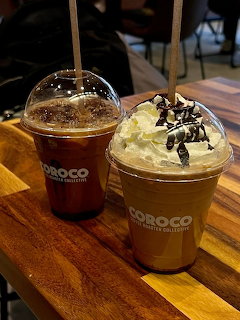Two iced coffee drinks in clear plastic cups on a wooden table, one with whipped cream topping