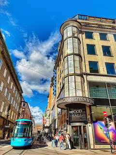 Blue tram on Oslo street with ornate corner building and pedestrians under sunny sky