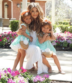 Mother hugging two young daughters in matching turquoise dresses with flower crowns in a flower garden