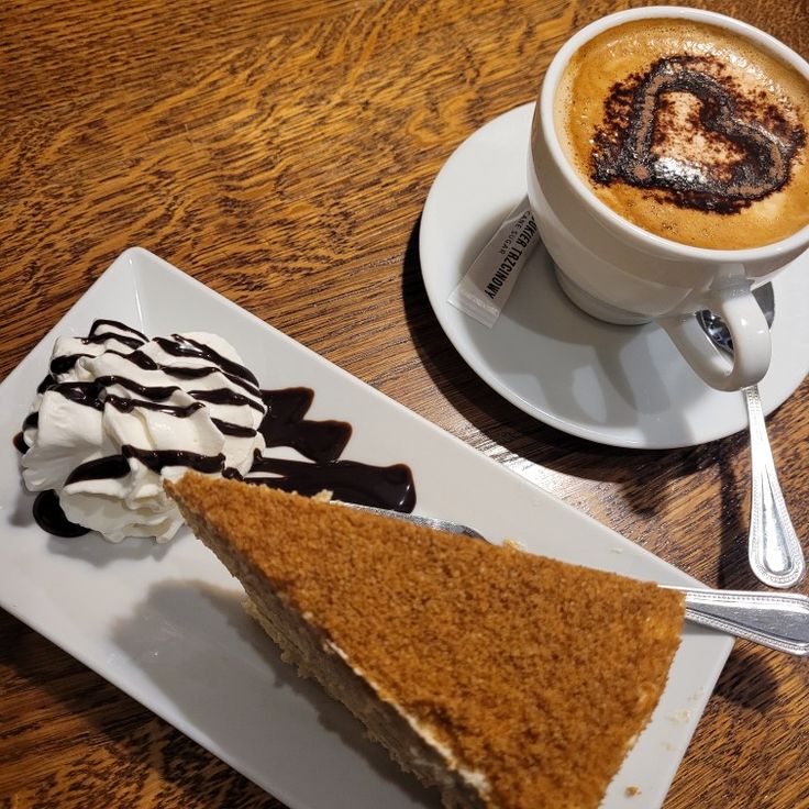 Slice of cake with chocolate sauce next to cappuccino with heart latte art on wooden table